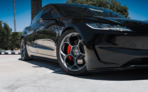 Front angle view of a 2024 Tesla Model 3 Performance in black, featuring 19-inch Satin Titanium UP-05 Forged Wheels by Unplugged Performance, parked under palm trees in a modern setting.