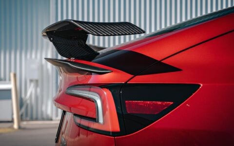 2024 Tesla Model 3 Performance in Ultra Red featuring a sleek carbon fiber wing spoiler by Unplugged Performance, parked in an industrial setting.
