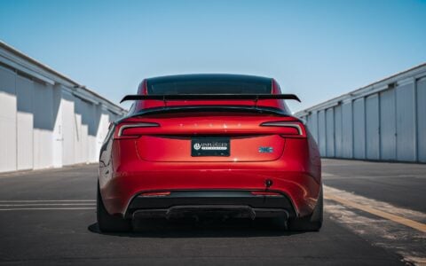 2024 Tesla Model 3 Performance in Ultra Red featuring a sleek carbon fiber wing spoiler by Unplugged Performance, parked in an industrial setting.