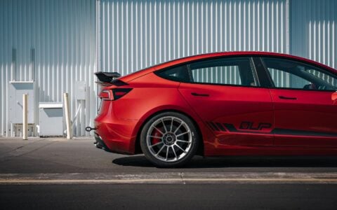 2024 Tesla Model 3 Performance in Ultra Red featuring a sleek carbon fiber wing spoiler by Unplugged Performance, parked in an industrial setting.