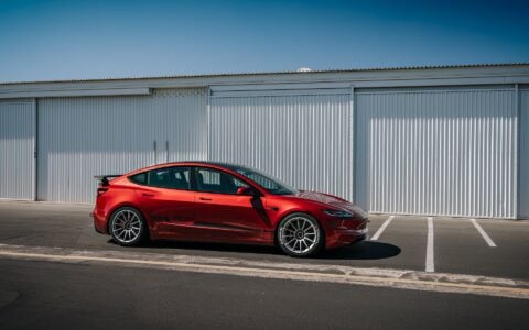 2024 Tesla Model 3 Performance in Ultra Red with UP-03 Pikes Peak Forged Wheels by Unplugged Performance, showcasing its aggressive stance and enhanced aerodynamics.; parked by an industrial setting.