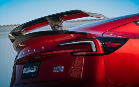 2024 Tesla Model 3 Performance in Ultra Red featuring a sleek carbon fiber wing spoiler by Unplugged Performance, parked in an industrial setting.
