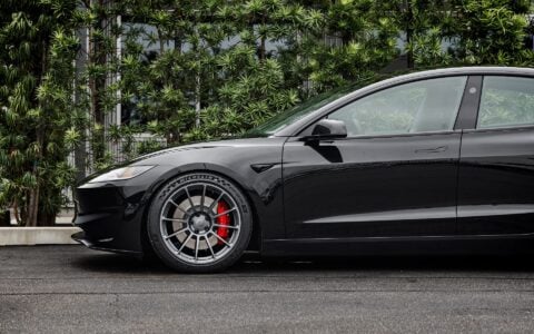 Side view of a 2024 black Tesla Model 3 Performance featuring Satin Titanium 19-inch UP-03 wheels by Unplugged Performance, parked in front of a modern building with green foliage and an overcast sky in the background.