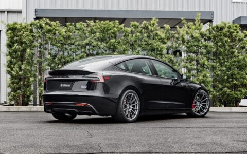 Rear side view of a 2024 black Tesla Model 3 Performance featuring Satin Titanium 19-inch UP-03 wheels by Unplugged Performance, parked on a wet asphalt surface with green foliage and a modern building in the background.