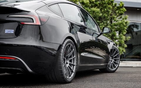 Rear side view of a 2024 black Tesla Model 3 Performance featuring Satin Titanium 19-inch UP-03 wheels by Unplugged Performance, parked on a wet asphalt surface with green foliage and a modern building in the background.