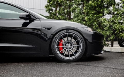 Front side profile of a 2024 black Tesla Model 3 Performance featuring Satin Titanium 19-inch UP-03 wheels by Unplugged Performance, parked in front of a modern building with green foliage in the background.