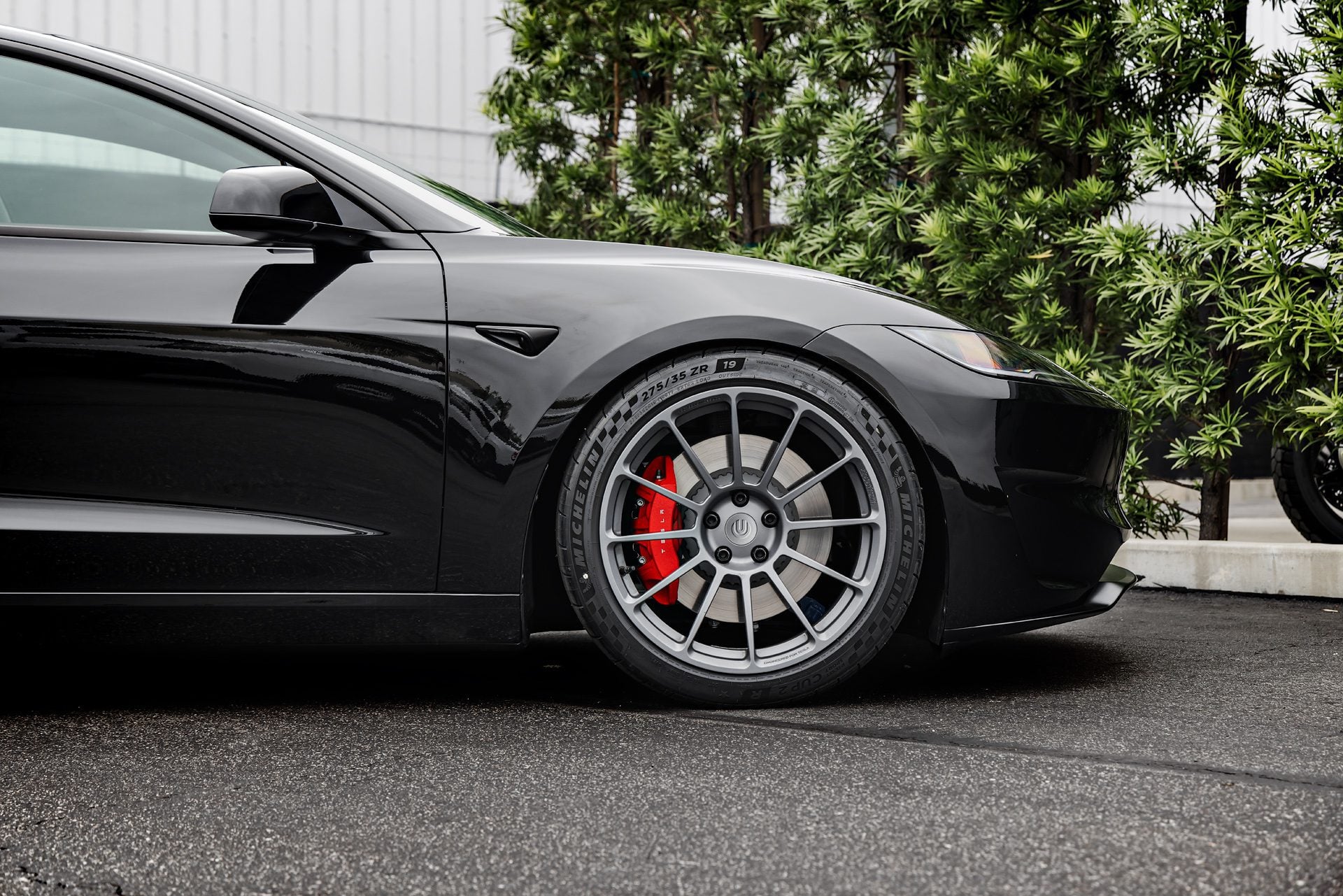 Front side profile of a 2024 black Tesla Model 3 Performance featuring Satin Titanium 19-inch UP-03 wheels by Unplugged Performance, parked in front of a modern building with green foliage in the background.