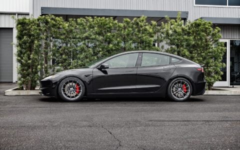 Side profile of a 2024 black Tesla Model 3 Performance featuring Satin Titanium 19-inch UP-03 wheels by Unplugged Performance, parked in front of a modern building with green foliage in the background.