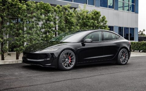 Front side view of a 2024 black Tesla Model 3 Performance featuring Satin Titanium 19-inch UP-03 wheels by Unplugged Performance, parked in front of a modern building with green foliage and an overcast sky in the background.