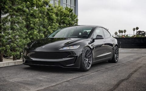 Front side view of a 2024 black Tesla Model 3 Performance featuring Satin Titanium 19-inch UP-03 wheels by Unplugged Performance, parked in front of a modern building with green foliage and an overcast sky in the background.