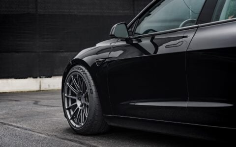 Front side view of a 2024 black Tesla Model 3 Performance featuring Satin Titanium 19-inch UP-03 wheels by Unplugged Performance, parked in front of a modern building with green foliage and an overcast sky in the background.