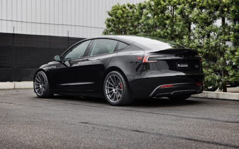 Rear side view of a 2024 black Tesla Model 3 Performance featuring Satin Titanium 19-inch UP-03 wheels by Unplugged Performance, parked in front of a modern building with green foliage and an overcast sky in the background.