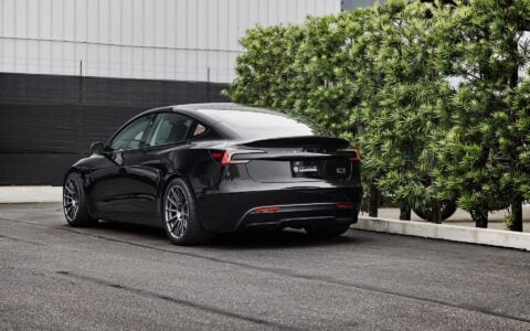 Rear side view of a 2024 black Tesla Model 3 Performance featuring Satin Titanium 19-inch UP-03 wheels by Unplugged Performance, parked in front of a modern building with green foliage and an overcast sky in the background.