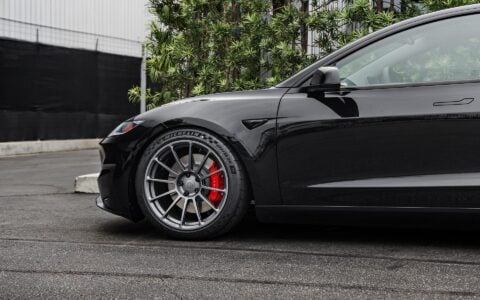Front side view of a 2024 black Tesla Model 3 Performance featuring Satin Titanium 19-inch UP-03 wheels by Unplugged Performance, parked in front of a modern building with green foliage and an overcast sky in the background.