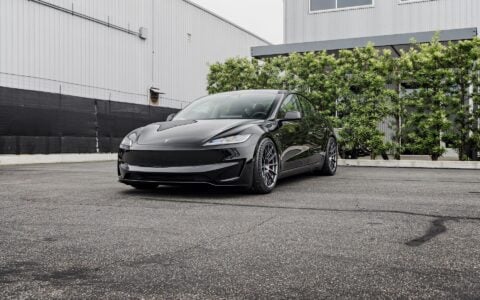 Front side view of a 2024 black Tesla Model 3 Performance featuring Satin Titanium 19-inch UP-03 wheels by Unplugged Performance, parked in front of a modern building with green foliage and an overcast sky in the background.