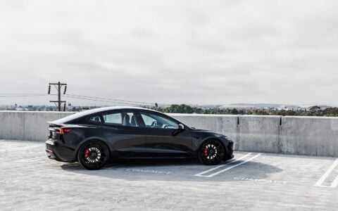 Rear side view of a black Tesla Model 3 Performance featuring 21-inch Gloss Black UP-03 wheels and red brake calipers by Unplugged Performance, parked in a rooftop parking lot under an overcast sky.