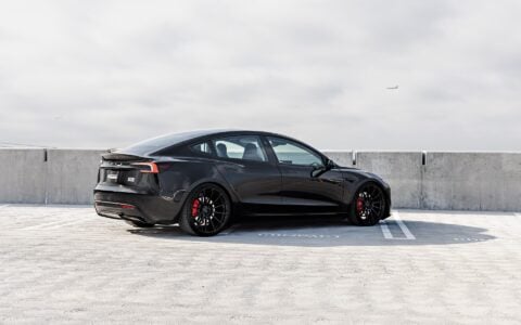 Rear side view of a black Tesla Model 3 Performance featuring 21-inch Gloss Black UP-03 wheels and red brake calipers by Unplugged Performance, parked in a rooftop parking lot under an overcast sky.
