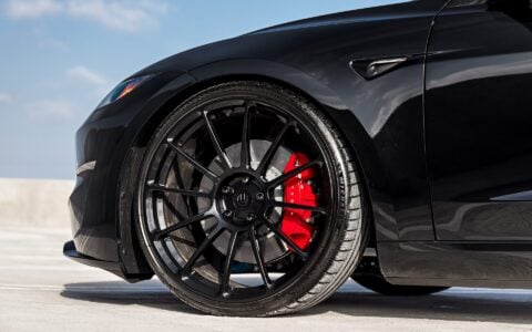 Close-up view of the front wheel of a black Tesla Model 3 Performance featuring 21-inch Gloss Black UP-03 wheels and red brake calipers by Unplugged Performance, parked on a rooftop parking lot under a clear sky.