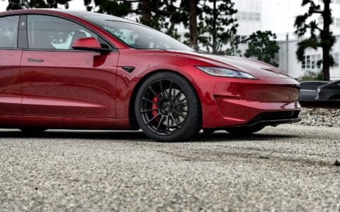 Angled front view of an Ultra Red Tesla Model 3 Performance with 19-inch Gloss Black UP-03 forged wheels by Unplugged Performance, parked near a train track background.