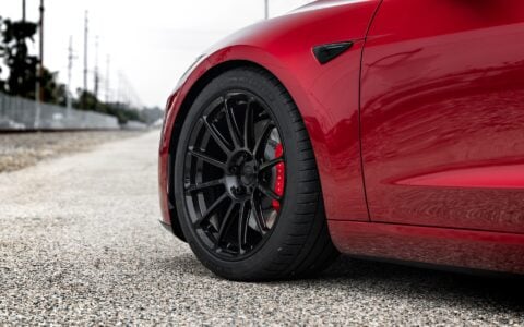 Close-up of the front wheel of an Ultra Red Tesla Model 3 Performance with 19-inch Gloss Black UP-03 forged wheels by Unplugged Performance, highlighting the wheel design and red brake calipers.