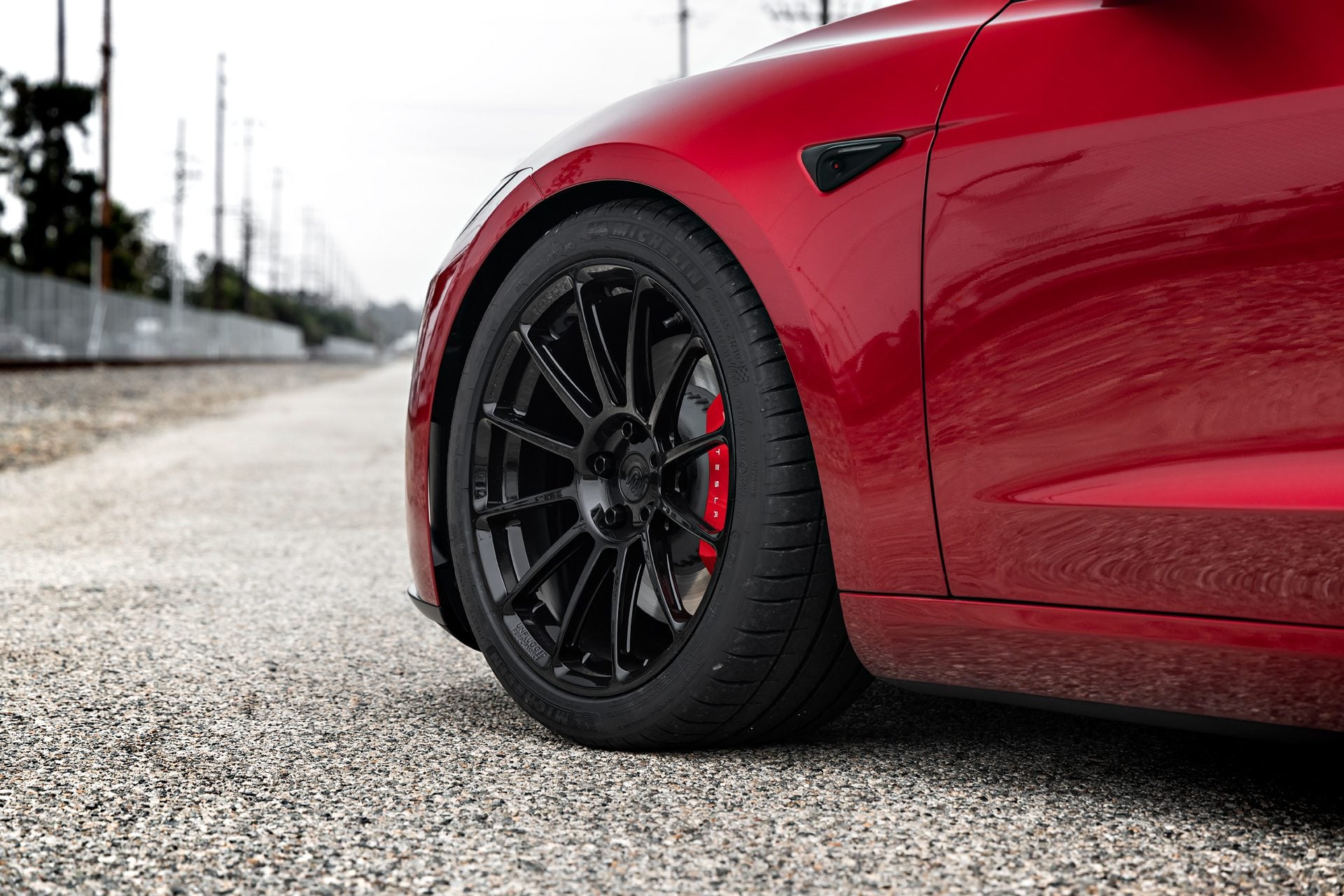 Close-up of the front wheel of an Ultra Red Tesla Model 3 Performance with 19-inch Gloss Black UP-03 forged wheels by Unplugged Performance, highlighting the wheel design and red brake calipers.