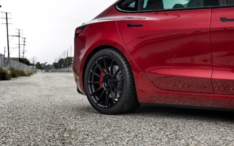 Rear quarter view of an Ultra Red Tesla Model 3 Performance featuring 19-inch Gloss Black UP-03 forged wheels by Unplugged Performance, parked on a gravel road near train tracks.