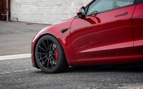 Angled front view of an Ultra Red Tesla Model 3 Performance showcasing 19-inch Gloss Black UP-03 forged wheels by Unplugged Performance, parked on an urban street.