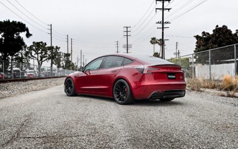 Rear side view of an Ultra Red Tesla Model 3 Performance with 19-inch Gloss Black UP-03 forged wheels by Unplugged Performance, parked in a lot with a black brick wall background.