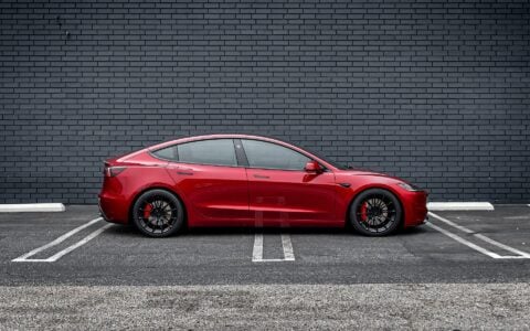 Side profile view of an Ultra Red Tesla Model 3 Performance equipped with 19-inch Gloss Black UP-03 forged wheels by Unplugged Performance, parked against a black brick wall.