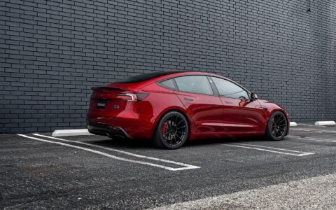 Rear profile view of an Ultra Red Tesla Model 3 Performance equipped with 19-inch Gloss Black UP-03 forged wheels by Unplugged Performance, parked against a black brick wall.