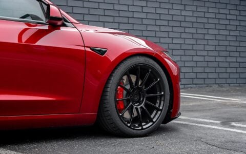 Front wheel view of an Ultra Red Tesla Model 3 Performance equipped with 19-inch Gloss Black UP-03 forged wheels by Unplugged Performance, parked against a black brick wall.