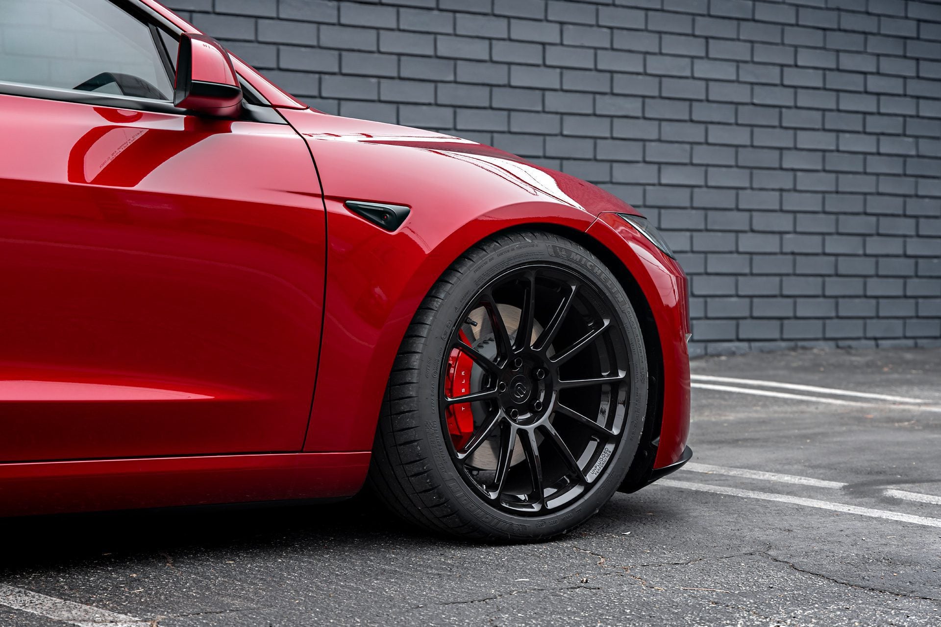 Front wheel view of an Ultra Red Tesla Model 3 Performance equipped with 19-inch Gloss Black UP-03 forged wheels by Unplugged Performance, parked against a black brick wall.