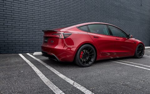 Profile view of an Ultra Red Tesla Model 3 Performance equipped with 19-inch Gloss Black UP-03 forged wheels by Unplugged Performance, parked against a black brick wall.