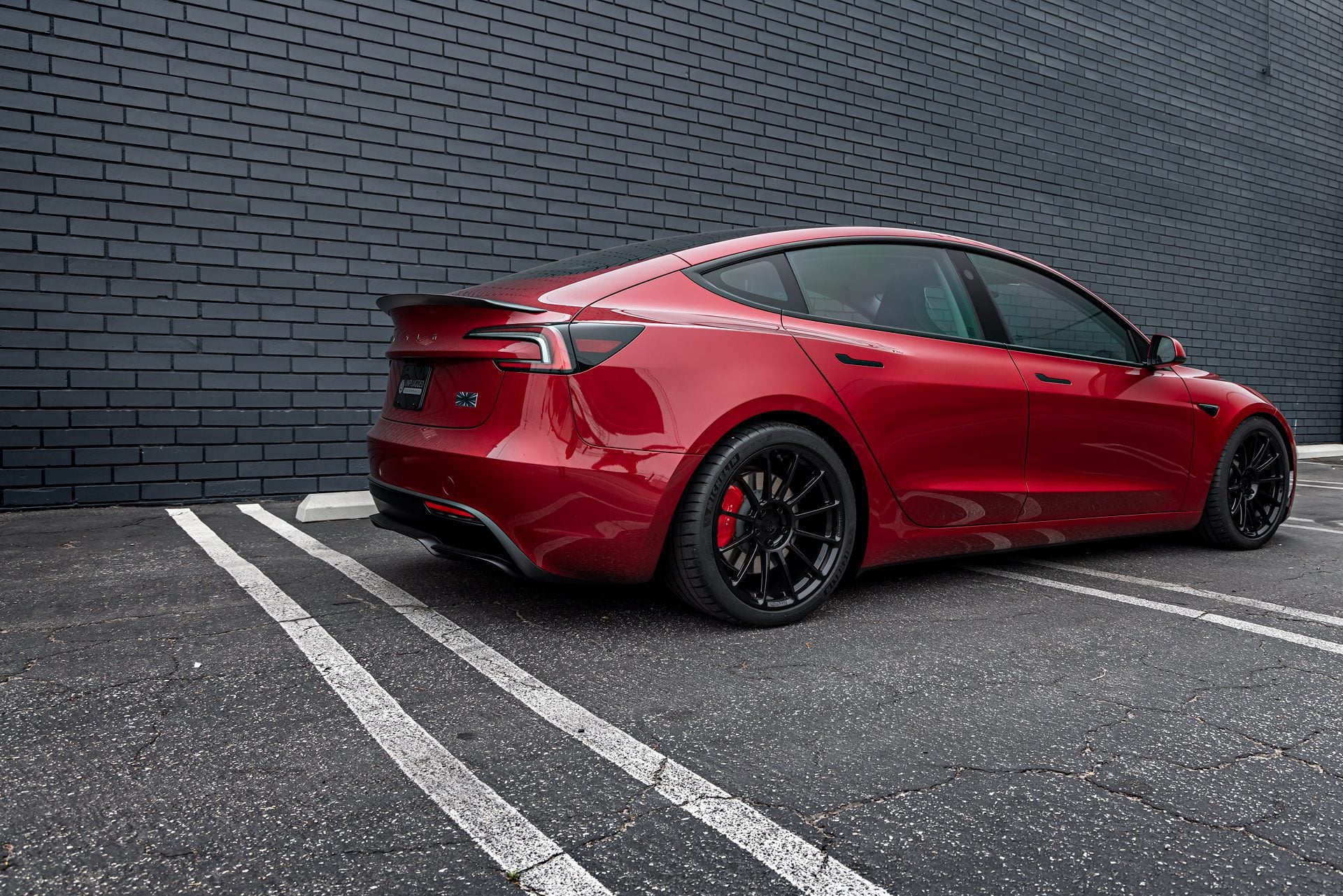 Profile view of an Ultra Red Tesla Model 3 Performance equipped with 19-inch Gloss Black UP-03 forged wheels by Unplugged Performance, parked against a black brick wall.
