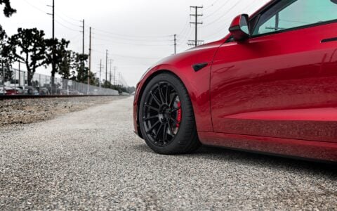 Angled front view of an Ultra Red Tesla Model 3 Performance with 19-inch Gloss Black UP-03 forged wheels by Unplugged Performance, parked near a train track background.