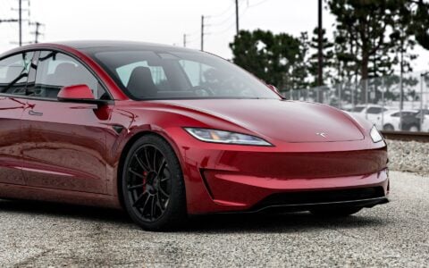 Angled front view of an Ultra Red Tesla Model 3 Performance with 19-inch Gloss Black UP-03 forged wheels by Unplugged Performance, parked near a train track background.