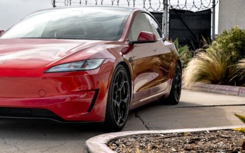 The image presents an Ultra Red Tesla Model 3 Performance from a front and side perspective, accentuating its 20-inch Gloss Black UP-05 Forged Wheels. The photo emphasizes the vehicle's dynamic stance and the wheels' premium finish.