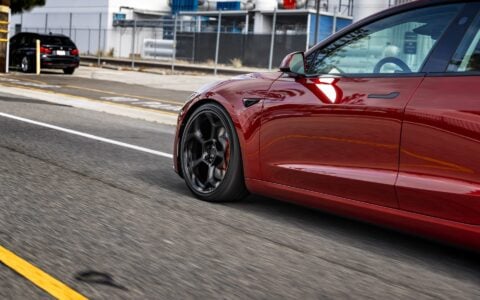 A rolling view of an Ultra Red Tesla Model 3 Performance, emphasizing its 20-inch Gloss Black UP-05 Forged Wheels. The photo captures the motion and elegance of the car, along with the distinctive wheel design.