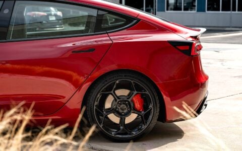 A close-up shot of the rear wheel of an Ultra Red Tesla Model 3 Performance, featuring the 20-inch Gloss Black UP-05 Forged Wheel from Unplugged Performance. The image highlights the wheel's detailed craftsmanship and the car's vibrant paintwork.